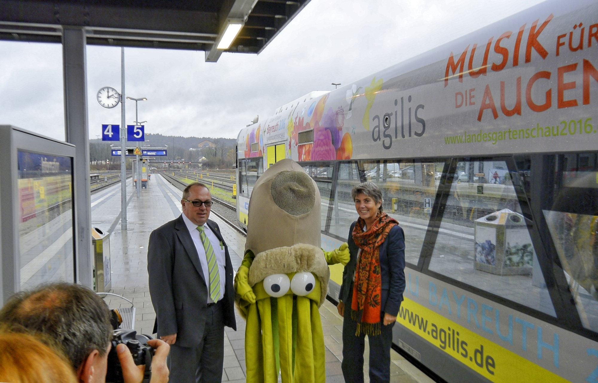 Vor dem agilis Zug mit der Werbung fr die Landesgartenschau 2016 in Bayreuth hieen der Geschftsfhrer der agils, Herr Dietmar Knerr, Frau Oberbrgermeisterin Brigitte Merk-Erbe und das tierische Maskottchen "Glotzaugen-Gerch" die Fahrgste des Zuges nach Weidenberg willkommen. Neben dem Logo unserer Festspiel- und Universittsstadt Bayreuth ist auf dem farbenfrohen Zug noch die Aussage der Landesgartenschau 2016 in Bayreuth angebracht: Musik fr die Augen. Im Hintergrund, leider nur schwach zu sehen, ist das berhmte Festspielhaus von Richard Wagner zu sehen. Denn mit Musik ist Bayreuth schon einmal weltberhmt geworden. Die Vorfreude auf den 22. April 2016 ist sehr gro. 