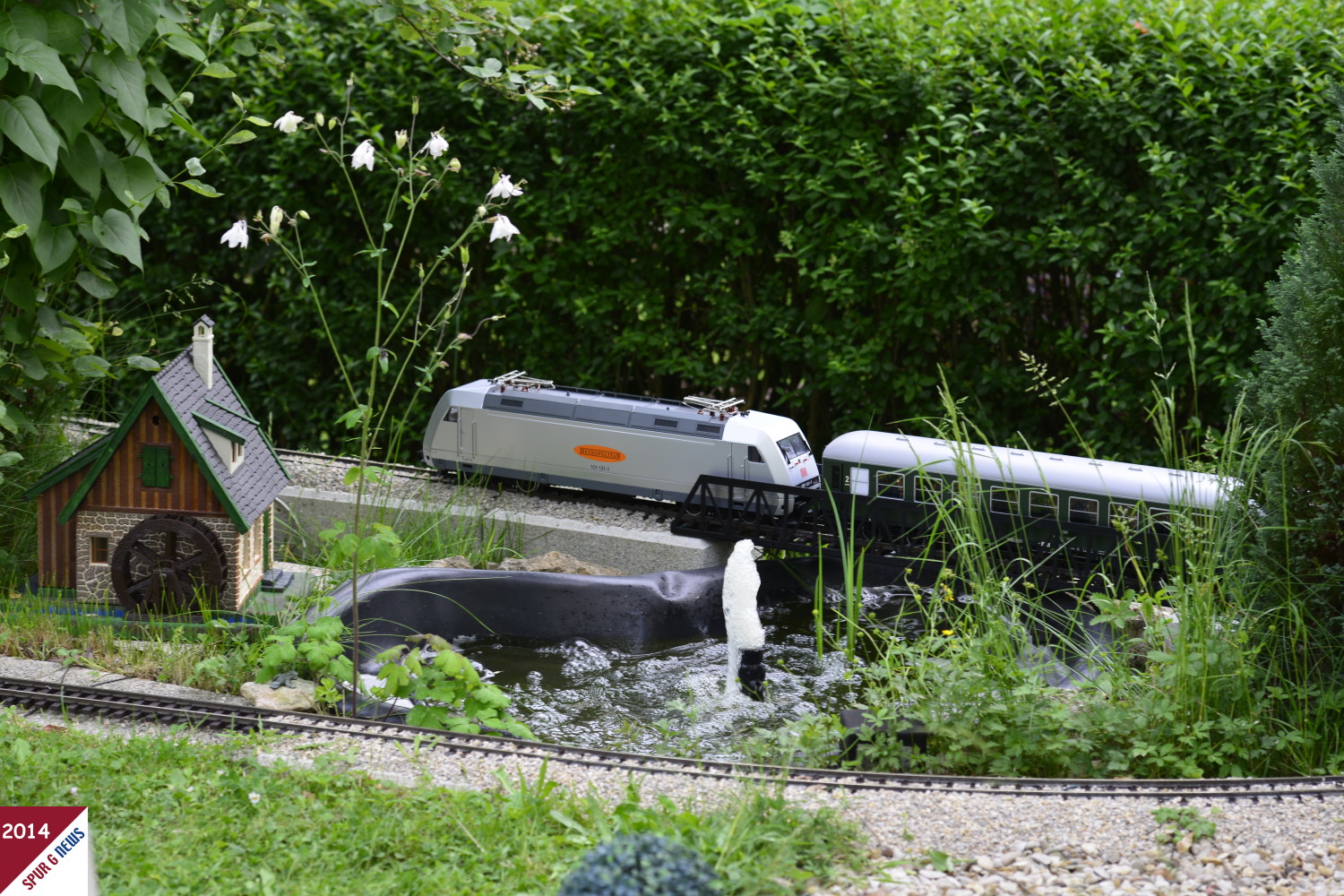 Eine DB 101 - Metropolitan 101 131-1 - zieht den Sonderzug, bestehend aus drei langen DB Reiszugwagen von LGB ber die Brcke am Mhlenteich der Erabocher-Gartenbahn. Idyllische Streckenfhrung an der Buchshecke ber die Gitterbrcke aus Metall hin zu ersten Schleife. Im Vordergrund der Streckenabschnitt der den Teich an der Mhle oberhalb passiert.