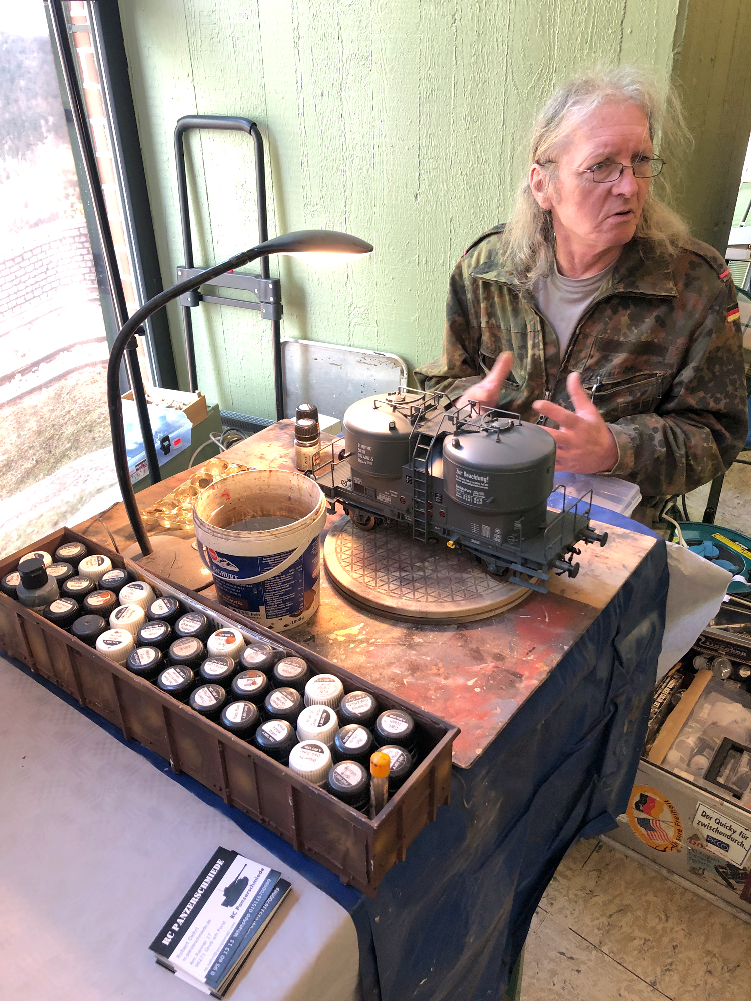 Das "Atelier" des Künstler mit - himself - Robert "Der Künstler". Einfach aufgebaut! Eine Holzplatte - so breit wie ein Biergartengarniturtisch, darauf eine Lampe, der Wassereimer zum Pinselwaschen und der Drehteller mit dem zu alternden Zementsilowagen. Am unteren Ende stehen die Farben bereit. Ein alter Hochbordwagen dient zur Aufbewahrung und zum einfachen Transport.