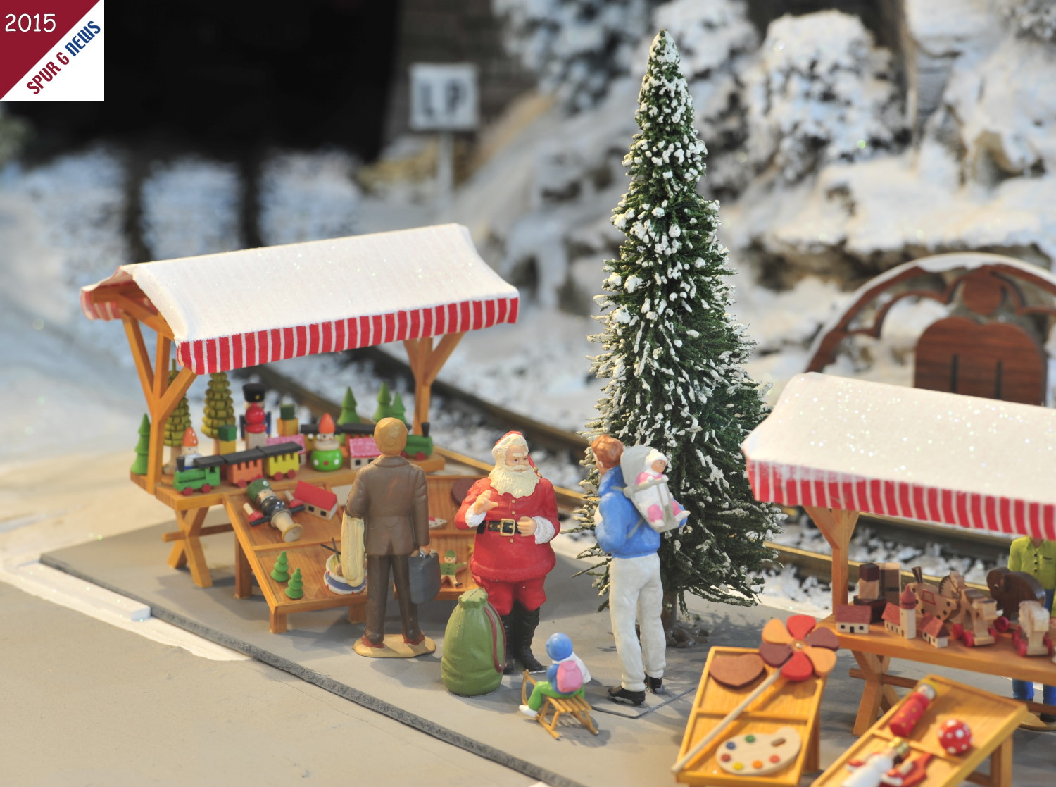 Das Erzgebirge mit der Erzgebirgischen Volkskunst liegen in Sachsen. Deshalb ist auch auf dieser Winteranlage von LGB ein typischer Weihnachtsmarkt mit Holzwaren, vom gedrehten Weihnachtsbaum ber die Erzengel bis hin zur Holzeisenbahn vorhanden. Wir wissen nicht, was der freundliche Nikolaus (Weihnachtsmann) dem Vater mit den beiden Kindern empfiehlt. Auf dem einen Marktstand steht eine Gartenbahn. Es wre schn, wenn diese Modellbahnen fr den Garten dem jungen Vater empfohlen werden. Natrlich knnte es auch sein, da der Sack vom Weihnachtsmann schon gefllt ist, die Wnsche der Kinder nach einer Eisenbahn erfllt sind