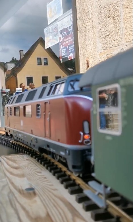 Kurzvideo von der Fahrt der Gartenbahnzüge vor dem Modellbaustudio Raab am 26. Juli 2025. Hier die V200 von LGB mit DB Wagen am Haken. In der zweiten Videohälfte ist eine Dampflok "Stainz" mit Personenwagen und zwei Güterwagen auf dem Rundkurs zu sehen. Im Schaufenster spiegeln sich zwar dunkle Wolken, jedoch konnte der Tag bei fast trockenem Wetter genossen werden.