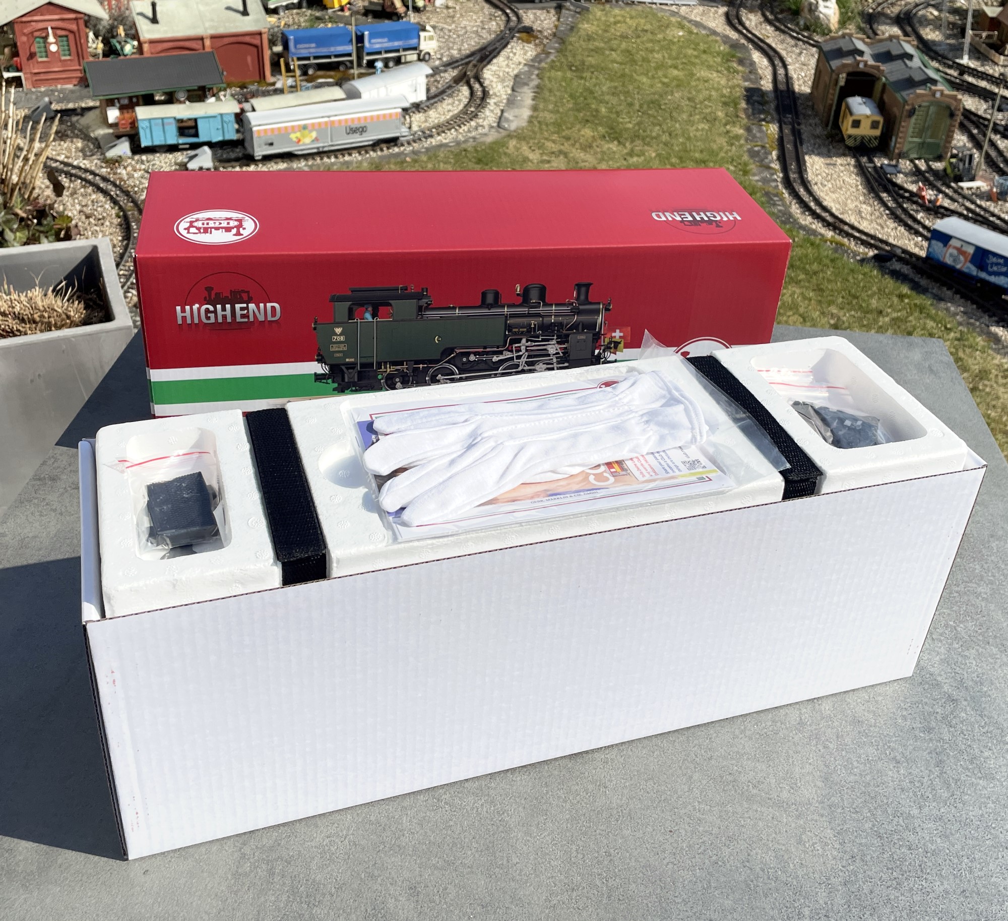 Der obere Teil des zweischaligen Verpackungskartons - rotes Oberteil - weißes Innenteil - gibt den Blick auf die feste Styroporverpackung frei. Wer schon mal eine Highend Metalllok, z.B. Krokodil ausgepackt hat, kennt den Anblick mit den beiden Klettbändern. Links und rechts sind in den Kartonvertiefungen neben Magneten, Maulschlüssel, Fahnen auch noch Kupplungen untergebracht. Im Mittelteil liegen die Baumwollhandschuhe, zur Vermeidung von Fingerabdrücken auf der Lok, bei. Darunter das Zertifikat, der Achslenker für die Weichen, die Bedienungsanleitung und die Pipette zum Einfüllen den Dampfloköls in den bereits werkseitig eingebauten Verdampfer.