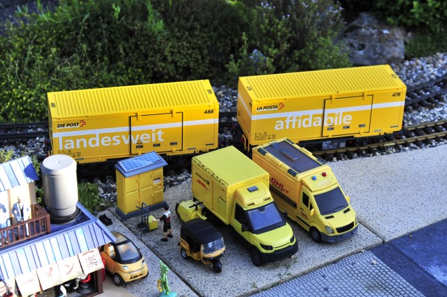 Spur G - Art.Nr. 45886. Auf der Anlage in Hummeltal fanden die beiden Postcontainerwagen bereits Ihren Platz am Paketlieferzentrum. Im Vordergrund ist eine Packstation mit angedeutetem Solarmodul, einem TukTuk von Temu, dem DHL Lieferfahrzeug von Simba Dickie und einem weiteren DHL Postfahrzeug zu sehen. Klar! Schweizer Postcontainer und DHL (deutsche Post) Fahrzeuge nebeneinander. Die DHL Fahrzeuge werden halt iin der Schweiz mal erprobt.