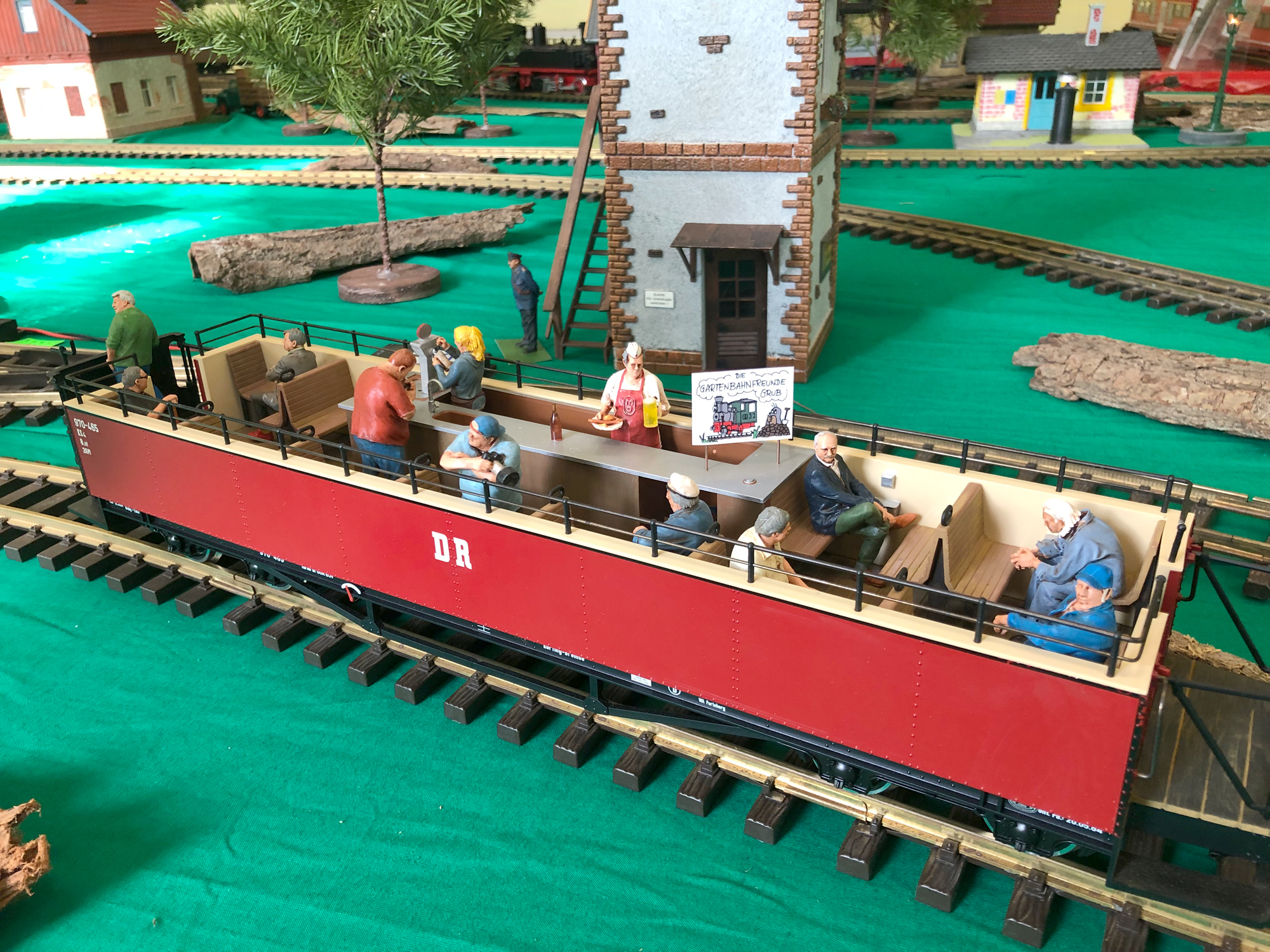 Der DR Cabriowagen mit der Bar und den Modellfiguren aus der Serie "Gartenbahnfreunde Grub a. Forst" mit Tresen, der Original Mitropa-Bedienung und der "Wirtin" darf nicht fehlen.