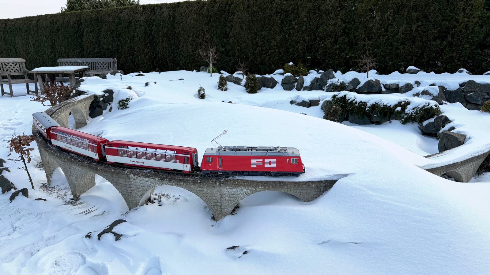 Das Modell der HGe 4/4 II der FO Nr. 101 - Art. Nr. 23103 von LGB - Herbstneuheit 2025 - mit dem kleinen Glacier Express auf der Gartenbahn im Norden mit Schneeverwehung. Es wird nun auf die Schneefräse gewartet.