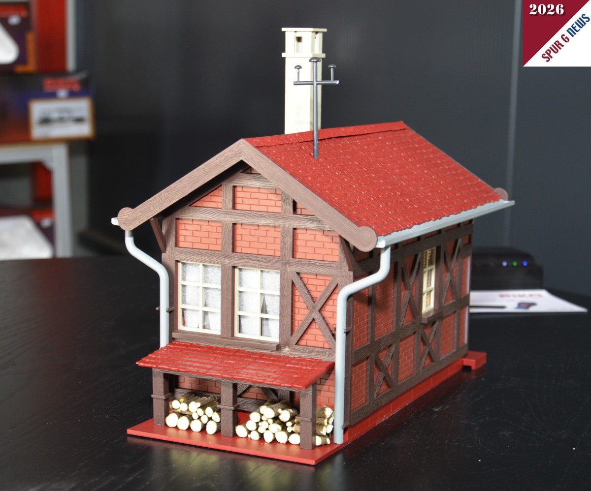 Das Bahnwärterhaus - Art. Nr. 65002 - konnten wir auf dem Präsentiertisch fotografieren. Link der Eingang des Hauses und rechts der Hinterhof mit dem gestapelten Holz unter der kleinen Überdachung. Sicherlich ragt der Kamin im Vorbild des Bahnwärterhauses auch so in die Höhe. Im Prospekt ist das Fachwerk weiß abgebildet. Hier beim Start in die Gartenbahnsaison sind es Klinkersteine die zwischen den aufgesetzten Balken eingebaut sind. Auch die Dachrinnen sind hier im Muster grau dargestellt. Im Prospekt grün. Wir werden erneut darüber berichten.
